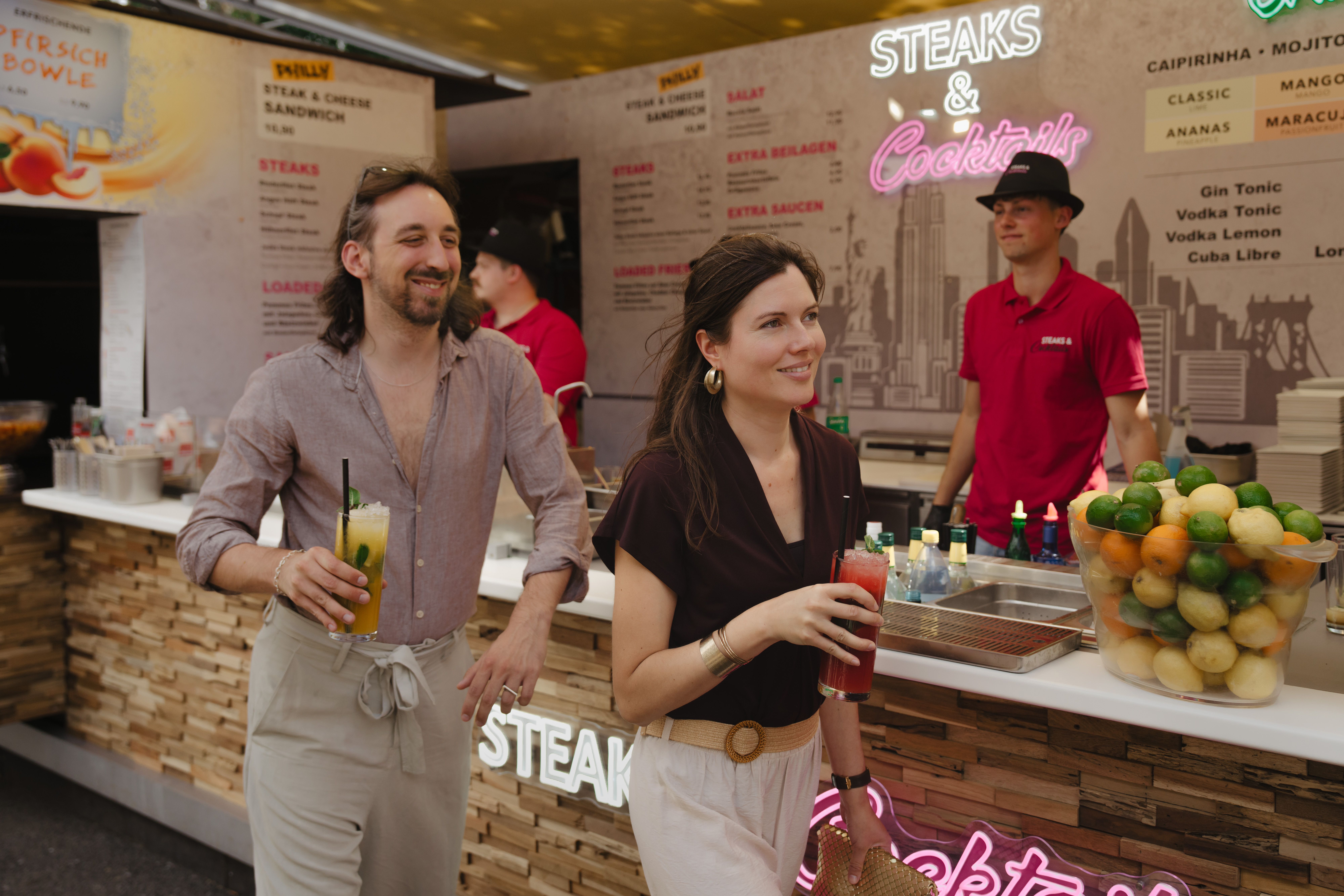 Zwei Personen vor einem Gastronomiestand mit zwei Getränken in der Hand