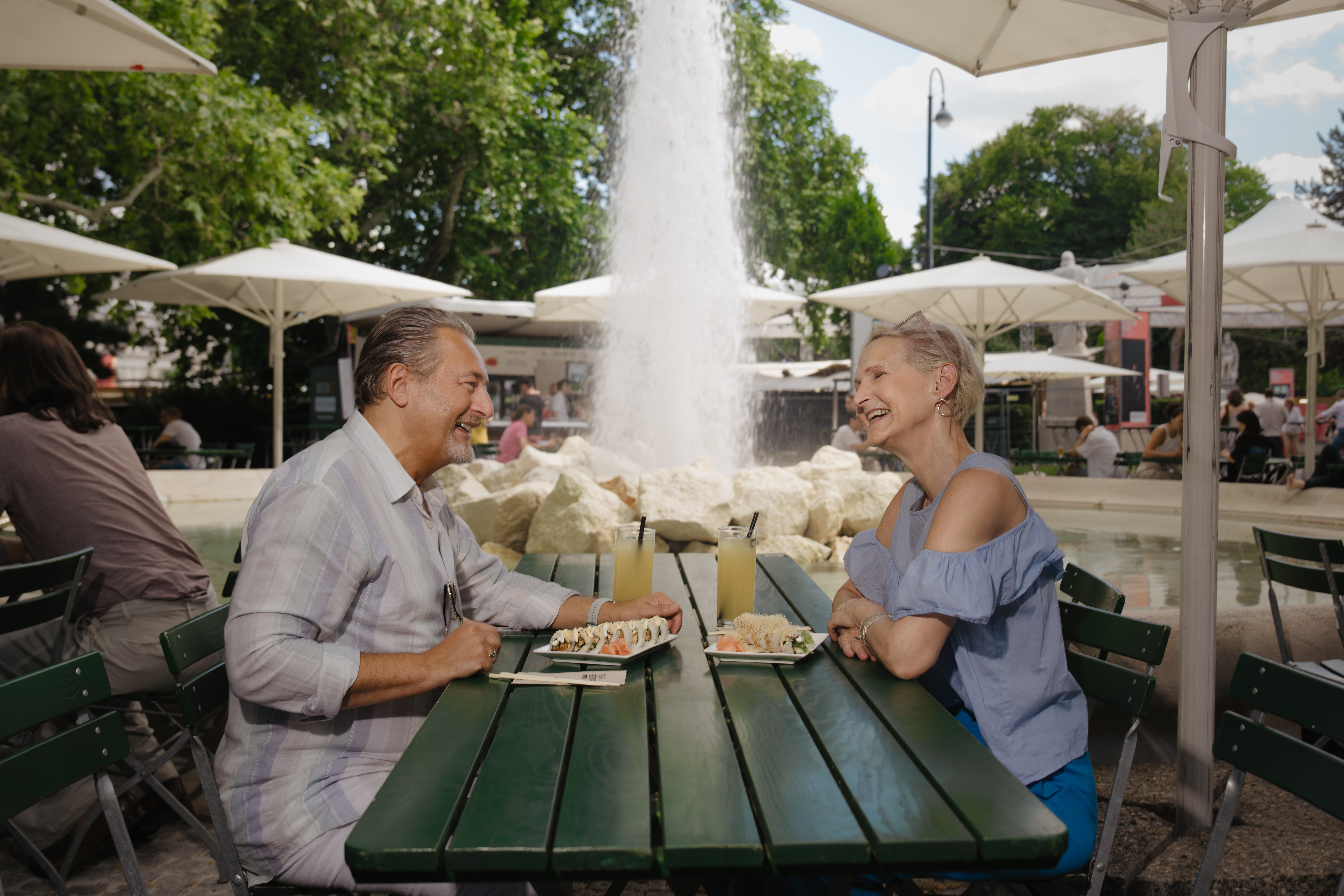 250628-FilmFestival-©flomitteregger-15 Ein Mann und eine Frau sitzen an einem Tisch und essen eine Speise. Im Hintergrund ist der Rathauspark zu sehen.