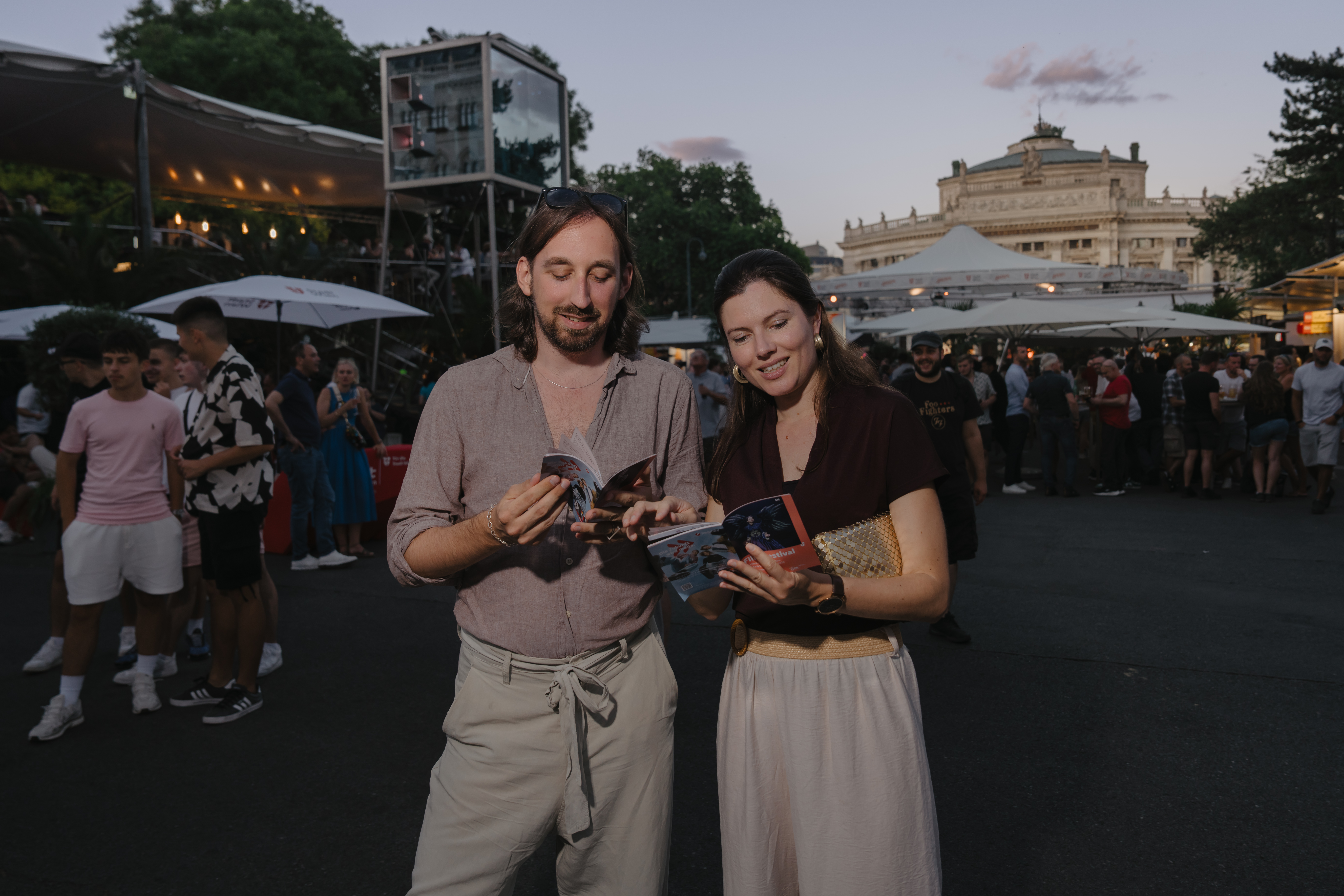 Ein Mann und eine Frau betrachten das Programm des Film Festivals. Sie stehen auf dem Festivalgelände, das im Hintergrund mit weiteren Besucherinnen und Besuchern sowie Festivalbereichen zu erkennen ist.