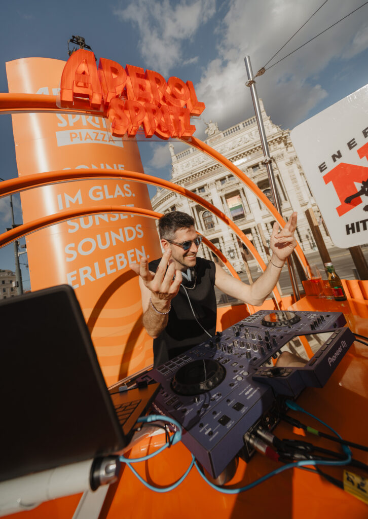 Auf dem Aperol-Stand steht ein DJ hinter seinem Mischpult und legt Musik auf.