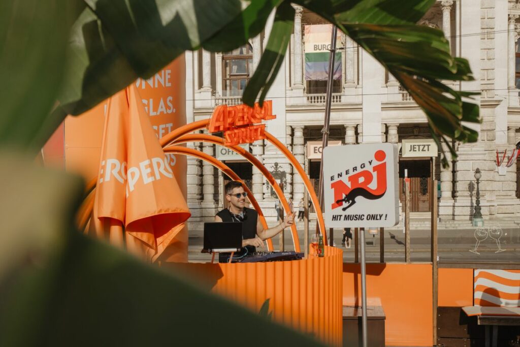 _250628-stadtwienmarketing-FilmFestival-flomitteregger-5 Kopie Auf dem Rathausplatz steht ein Aperol-Stand, vor dem sich mehrere Menschen aufhalten. Einige Besucher sitzen in der Nähe mit Getränken und genießen die Veranstaltung.