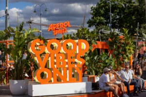 Auf dem Rathausplatz befindet sich ein Aperol-Stand. Daneben steht ein Aufsteller mit der Aufschrift ‚Good Vibes Online‘. Mehrere Menschen sitzen und stehen in der Nähe des Standes, einige mit Getränken. Die Atmosphäre wirkt entspannt und gesellig.